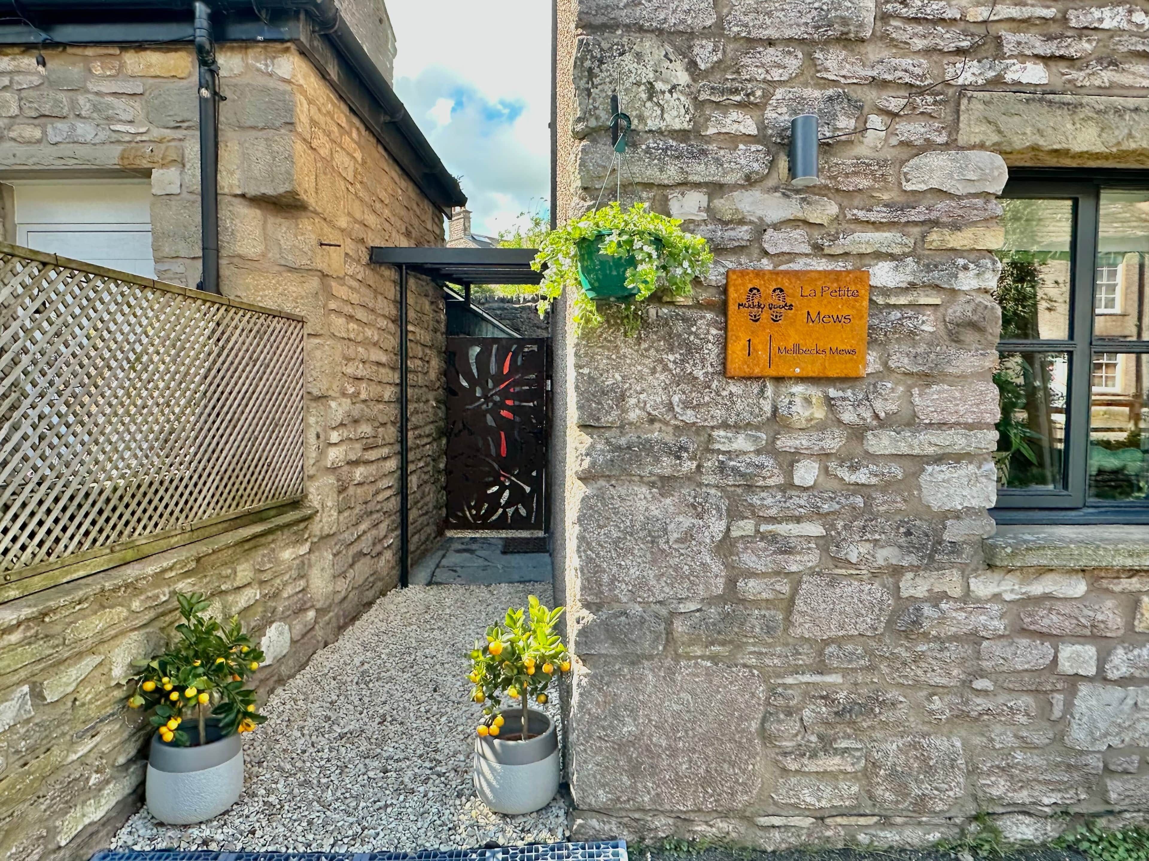 A stone building with a sign indicating 'La Petite Mews' and a number '1'. There are two potted plants in front.