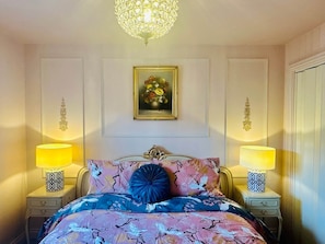 A bedroom with a bed, two bedside lamps, a chandelier, a framed floral painting, and a decorative wall panel. - Muddy Boots-La Petite Mews (Kirkby Stephen)