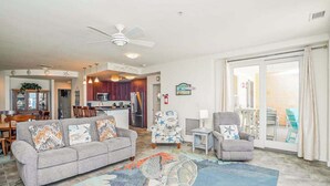 A living room with a sofa, armchairs, a ceiling fan, and a kitchen in the background. - Beachfront condo with 2 oceanfront balconies, W/D, pool, & gym (Virginia Beach)