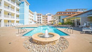 A swimming pool with a fountain, surrounded by lounge chairs and a multi-story building with balconies. - Comfortably furnished condo with glimpses of Back Bay, pool & gym (Virginia Beach)