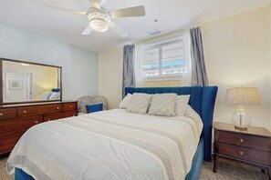 Une chambre à coucher comprenant un lit, une commode, une table de chevet, un ventilateur de plafond et une fenêtre avec des rideaux.