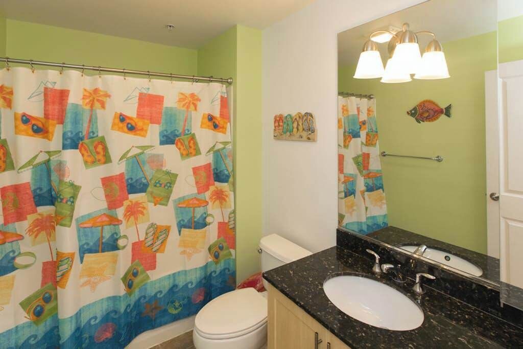 Un baño con cortina de ducha con diseño tropical, inodoro blanco, lavamanos con espejo y barra para toallas.