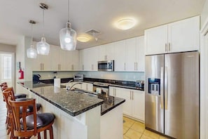 Eine moderne Küche mit weißen Schränken, einem Edelstahl-Kühlschrank, einer Mikrowelle und einer Kücheninsel mit Granitplatte und Barhocker.