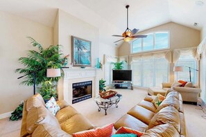 A living room with a fireplace, a television, a ceiling fan, and a large window with blinds. - Fantastic oceanfront home w/private pool & hot tub, WiFi, central AC, & deck (Virginia Beach)