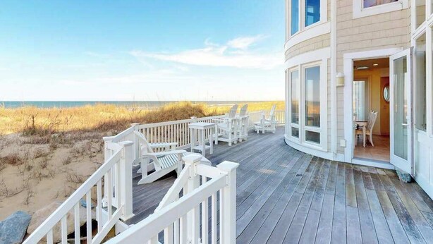 Een pand aan het strand met een terras, stoelen en grote ramen.