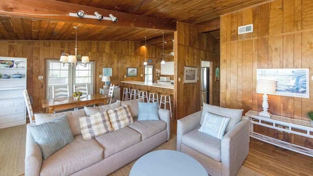 Ein gemütliches Wohnzimmer mit einer Couch, zwei Sesseln, einem Couchtisch, Holzwänden, einer Küchenzeile und einem Esstisch mit Barhocker.
