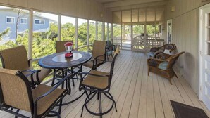 Eine überdachte Terrasse im Freien mit rundem Tisch und Stühlen, Blick auf ein Haus und Bäume sowie eine Holzterrasse.