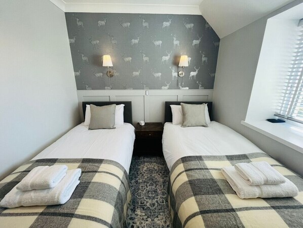 A twin room with two beds, a patterned wallpaper, bedside lamps, and a window with blinds.