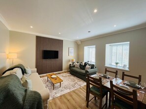 A living room with a sofa, a coffee table, a TV mounted on the wall, and a dining area with a table set for four.
