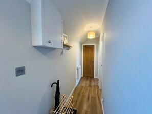 A narrow hallway with a wooden floor, a white wall, a door at the end, and a hanging rack with an umbrella.