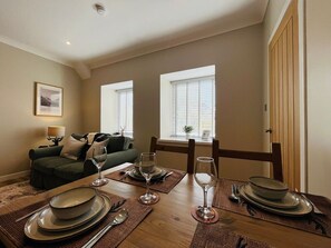A dining area with a wooden table set for four, a green sofa, and two windows with blinds. - Nevis Nook near Fort William (Torlundy)