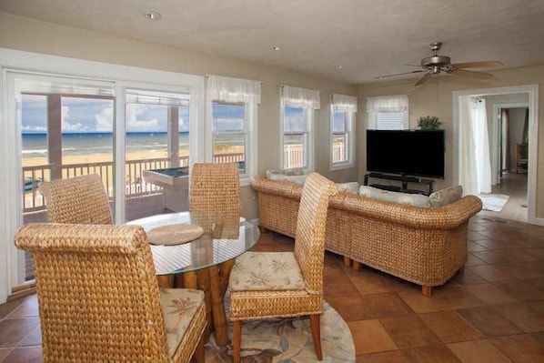 A living room with wicker furniture, a glass table, a flat-screen TV, and a view of the beach. - Oceanfront home with pool, hot tub, central AC, & furnished deck - near shopping (Virginia Beach)