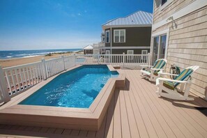 A pool on a wooden deck with a view of the beach, a house with a metal roof, and two lounge chairs. - Oceanfront home with pool, hot tub, central AC, & furnished deck - near shopping (Virginia Beach)