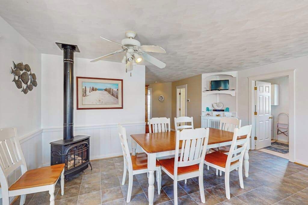 A dining area with a wooden table and chairs, a white ceiling fan, a black wood stove, and a framed picture on the wall.