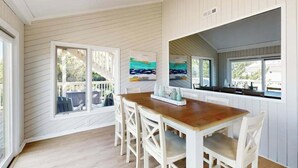 Una cocina con mesa de madera y sillas blancas, ventanas amplias y vistas al exterior.