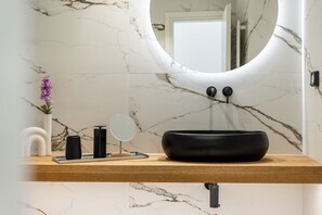 Un bany amb paret de marbre, un mirall rodó, un lavabo negre i un taulell de fusta.