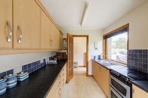 A kitchen with wooden cabinets, a black countertop, a stove, and a window with a view of a car outside. - Tucked away cottage with fantastic views in the Cairngorms national park. (Inveruglass)