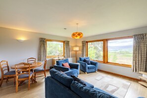 A living room with a sofa, chairs, a dining table, and two windows with curtains. - Tucked away cottage with fantastic views in the Cairngorms national park. (Inveruglass)