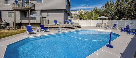 A rectangular swimming pool with blue water, surrounded by a white deck. There are blue lounge chairs and a white fence in the background.
