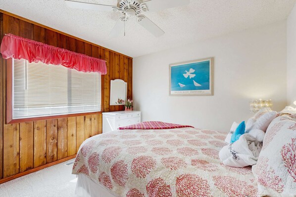 Ein Schlafzimmer mit einer holzverkleideten Wand, einem weißen Deckenventilator, einem Fenster mit Jalousien, einem weißen Schminktisch und einem Bett mit gemusterten Betttextilien. An der Wand hängt ein gerahmtes Bild.