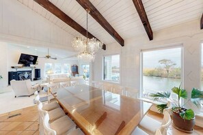 A spacious dining area with a long wooden table, white chairs, and a chandelier. There is a television and a fireplace in the background. - Attractive canal-front home w/private pool, WiFi, washer/dryer, central AC, deck (Virginia Beach)