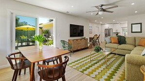 A modern living room with a dining area, a ceiling fan, a flat-screen TV, and a large window with an umbrella. - Coastal Palms - Walkable to Lower Village (Montecito)