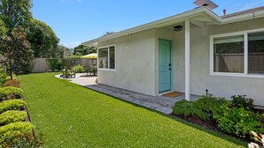 A single-story house with a green door, a patio with a table and chairs, and a well-maintained lawn. - Coastal Palms - Walkable to Lower Village (Montecito)
