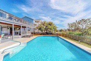 Ein Schwimmbecken mit Holzterrasse und Liegestühlen, umgeben von einem Zaun und Bäumen.