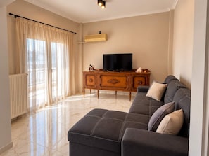 A living room with a sectional sofa, a television on a wooden cabinet, and a radiator. - Nea Ionia Urban Nest – Fotis’ Athens Getaway (Nea Ionia)