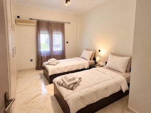 A hotel room with two beds, a window with curtains, a bedside table with a lamp, and a wall-mounted air conditioning unit. - Nea Ionia Urban Nest – Fotis’ Athens Getaway (Nea Ionia)