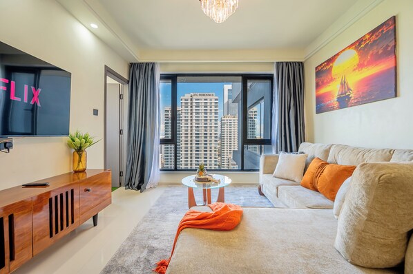 A modern living room with a sofa, a TV, a plant, and a view of high-rise buildings. - Misty Springs 1BR near GTC - Heated Infinity Pool, Gym, Fast WiFi (Nairobi)