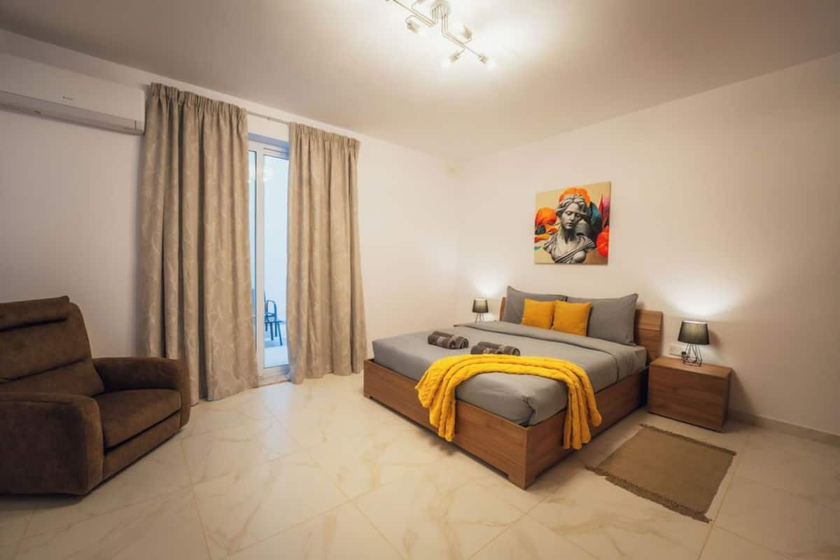 Un dormitorio con cama, una silla, una mesita de noche, un cuadro y una ventana con cortinas.
