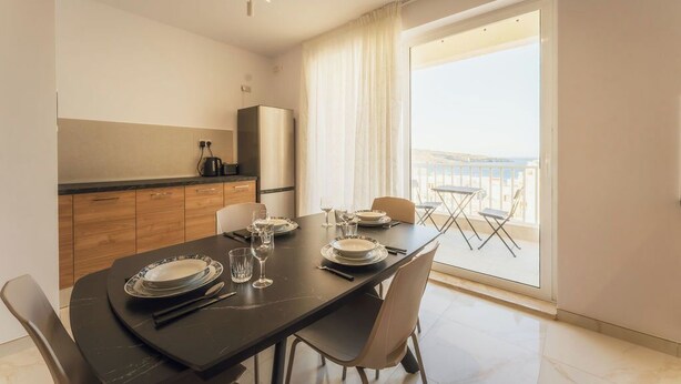 Un comedor con una mesa preparada para cuatro, una cocina con refrigerador y un balcón con vistas.