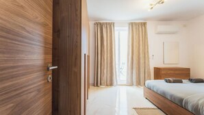 Un dormitorio con puerta de madera, cama con cabecera, mesita de noche, ventana con cortinas y aire acondicionado instalado en la pared.