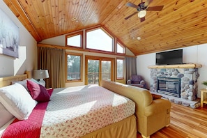 A bedroom with a large bed, a stone fireplace, a ceiling fan, and a TV mounted on the wall.