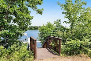A wooden bridge leading to a body of water, surrounded by dense greenery. - Rustic lakefront home with shared dock & boat slip, game room & stunning gazeebo (Necedah)