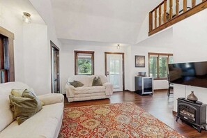 A spacious living room with a sofa, a patterned rug, a television, and a wood-burning stove. - Cozy Home in the trees, w/ large yard, fire pit, gas grill, & WIFI (Friendship)