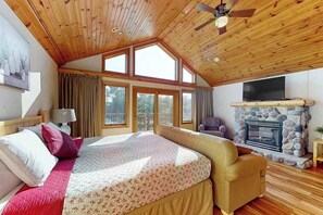 A bedroom with a large bed, a stone fireplace, a television, and a ceiling fan.