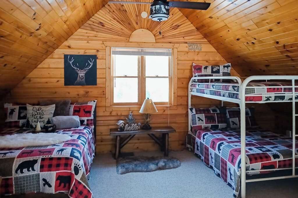Ein gemütliches Blockhaus mit Holzwänden, einem Satteldach, einem großen Fenster, Etagenbetten, einem Tisch mit Lampe und einer Bettdecke mit Reh-Motiv.