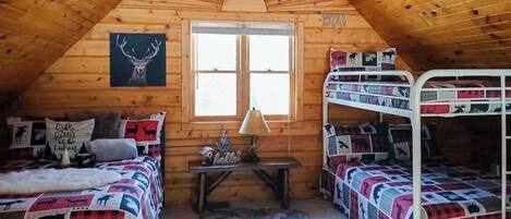 A cozy cabin with wooden walls, a pitched roof, a large window, bunk beds, a table with a lamp, and a deer-themed bedspread.