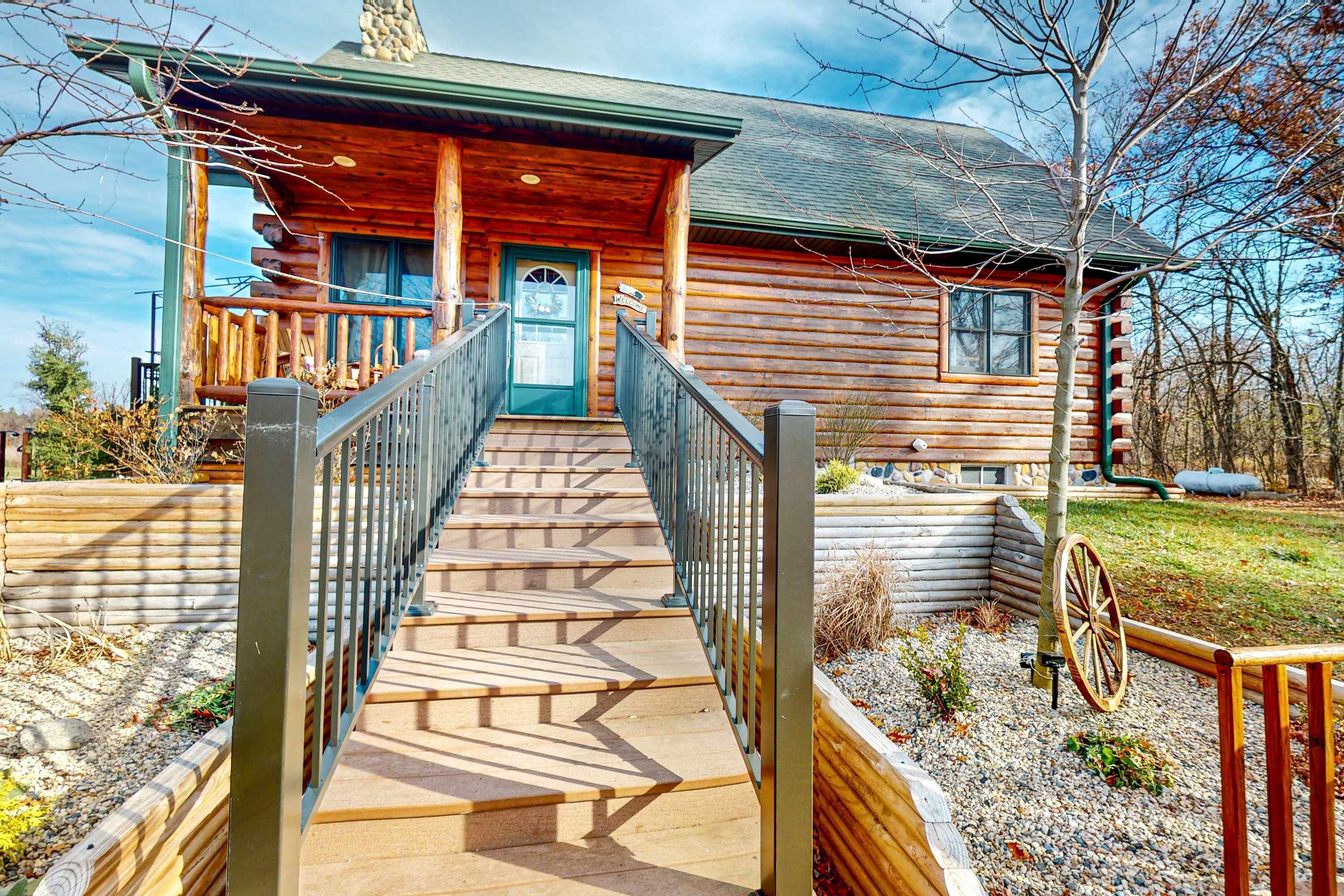 A log cabin with a wooden staircase leading to a front door, a porch with railings, and a wheelbarrow in the yard.
