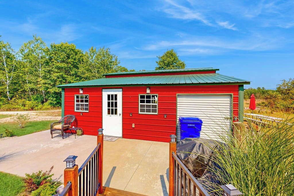A red single-story building with a green roof, a white garage door, and a small porch with a chair and table.