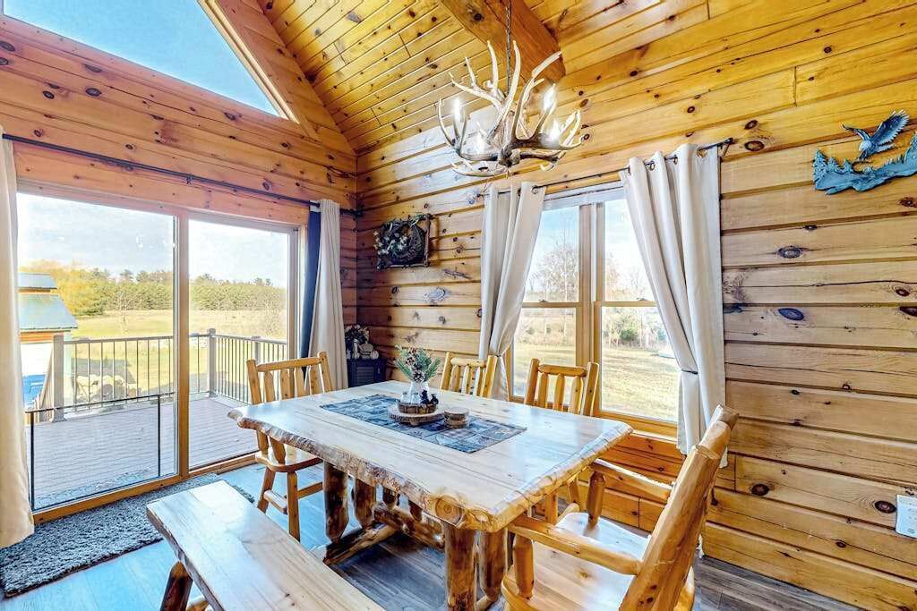 A wooden cabin interior with a dining table, chairs, a chandelier, and large windows.