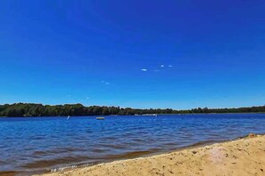 A beach with clear blue water, a sandy shore, and a forested area in the background. - Cozy Home in Lake Arrowhead w/ shared amenities such as pools, tennis, & golf (Nekoosa)