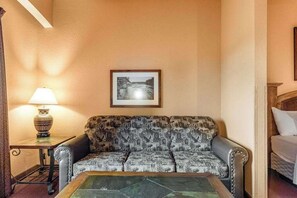 A room with a patterned sofa, a framed picture on the wall, a lamp on a side table, and a bed with white bedding.