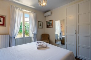 A hotel room with a bed, a chair, a window with curtains, a mirror, and wall-mounted air conditioning. - Casa Elisa - Lucca with Private Garden and Garage (Lucca)