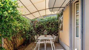 Eine überdachte Terrasse mit Tisch und Stühlen, umgeben von üppiger Bepflanzung und einem Spalier.