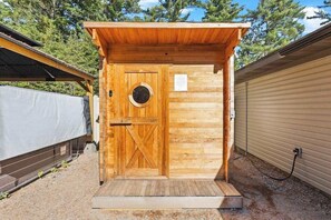 Estructura de sauna de madera con puerta, un pequeño ventanal circular y un escalón que lleva a ella.