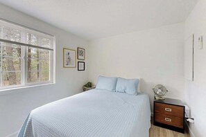 A bedroom with a bed, a bedside table with a fan, a nightstand, and a window with blinds.