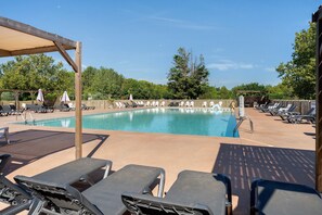 Un área de piscina con sillones de descanso y sombrillas, rodeada por una terraza de madera y árboles.